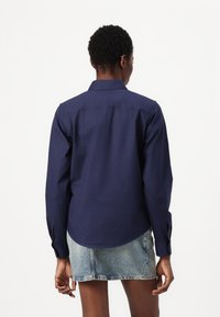 Camicia blu navy a maniche lunghe con colletto, orlo dritto e polsini abbottonati. Indossata con una mini gonna di denim chiaro. Texture semplice e liscia.