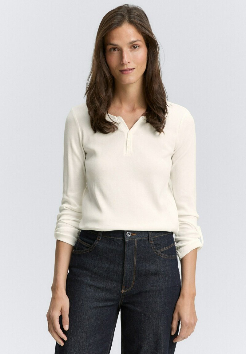 Giovane donna con lunghi capelli castani, che indossa una camicia bianca a maniche lunghe con bottoni e jeans blu scuro, in piedi su uno sfondo neutro.