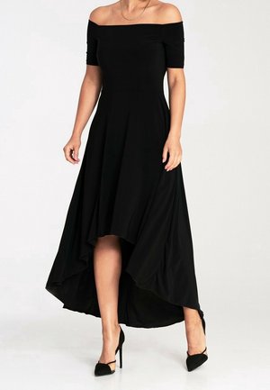 Femme portant une robe noire à épaules dénudées, courte devant et longue derrière, avec manches courtes et chaussures à talons hauts noires sur fond blanc uni.