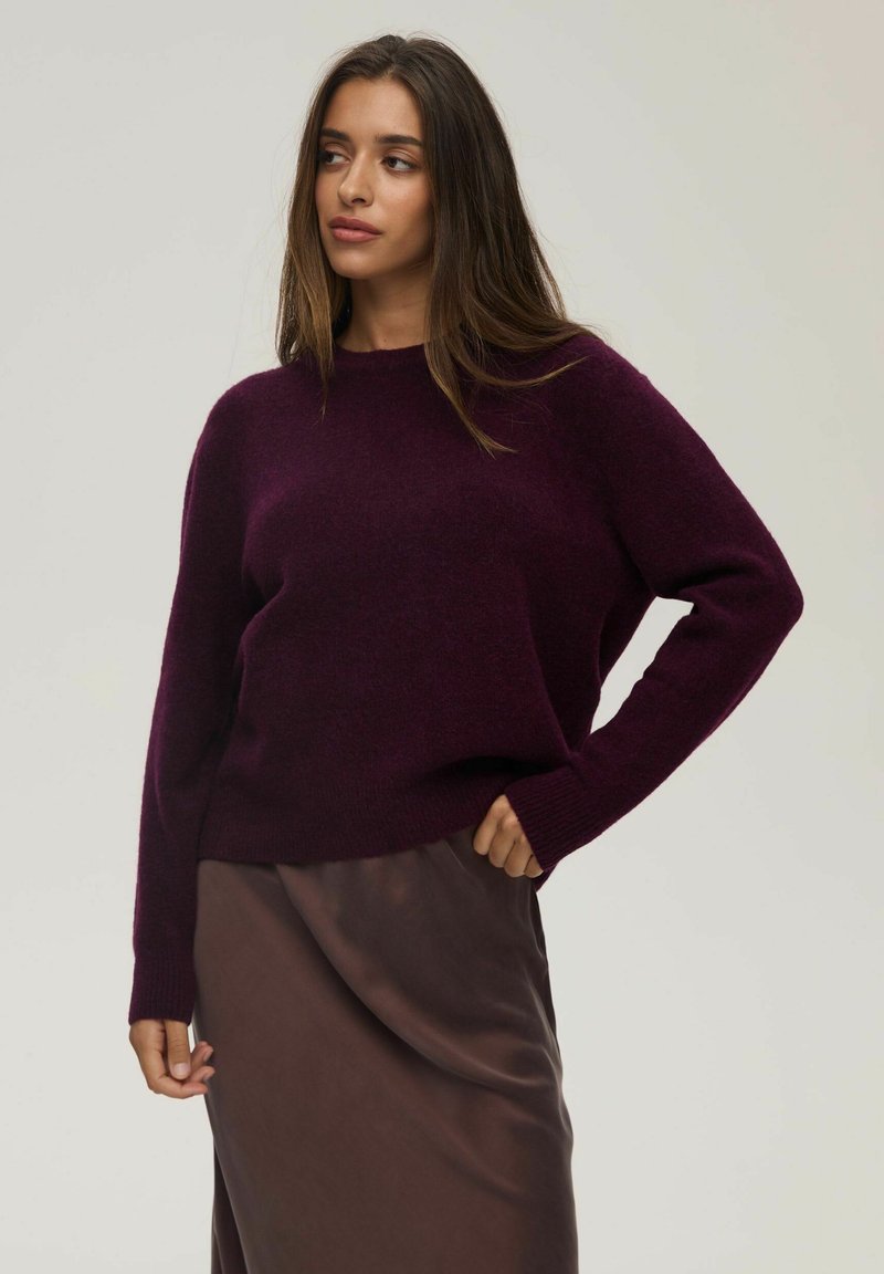 Burgundowa dziergana sweter z okrągłym dekoltem, opadającymi ramionami i ściągaczami na mankietach, w zestawieniu z brązową satynową spódnicą. Teksturowany, miękki materiał.