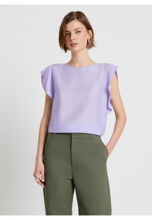 Frau trägt eine hellviolette ärmellose Bluse mit Rüschenärmeln und olivgrüne hochgeschnittene Hose, posiert mit einer Hand in der Tasche.