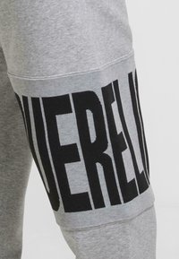 Šedé tepláky s veľkým čiernym textovým grafickým dizajnom "DERE" na boku, vyrobené z jemnej bavlnenej látky s hladkou textúrou.