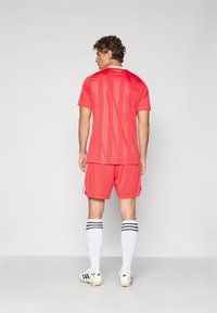 Rode sporttop met verticale strepen, gecombineerd met bijpassende shorts. Witte sokken met zwarte strepen en zwarte voetbalschoenen maken de outfit compleet.