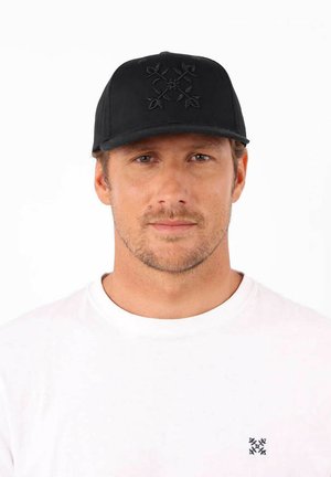 Casquette noire au design structuré, avec un emblème en relief sur le devant. Portée avec une chemise blanche simple, affichant un look décontracté.