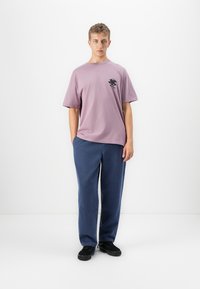 Noor mees seisab käed taskus, kandes heledat lilla üleküpset T-särki väikese musta graafikuga ning lahtisi siniseid pükse mustade kingadega.