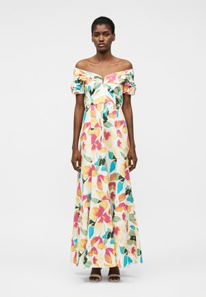 Γυναίκα που φοράει ένα μακρύ, φλοράλ φόρεμα με ανοιχτούς ώμους, με μοτίβα σε ροζ, κίτρινο, πράσινο και μπλε, στέκεται μπροστά σε ένα απλό ανοιχτό γκρι φόντο.