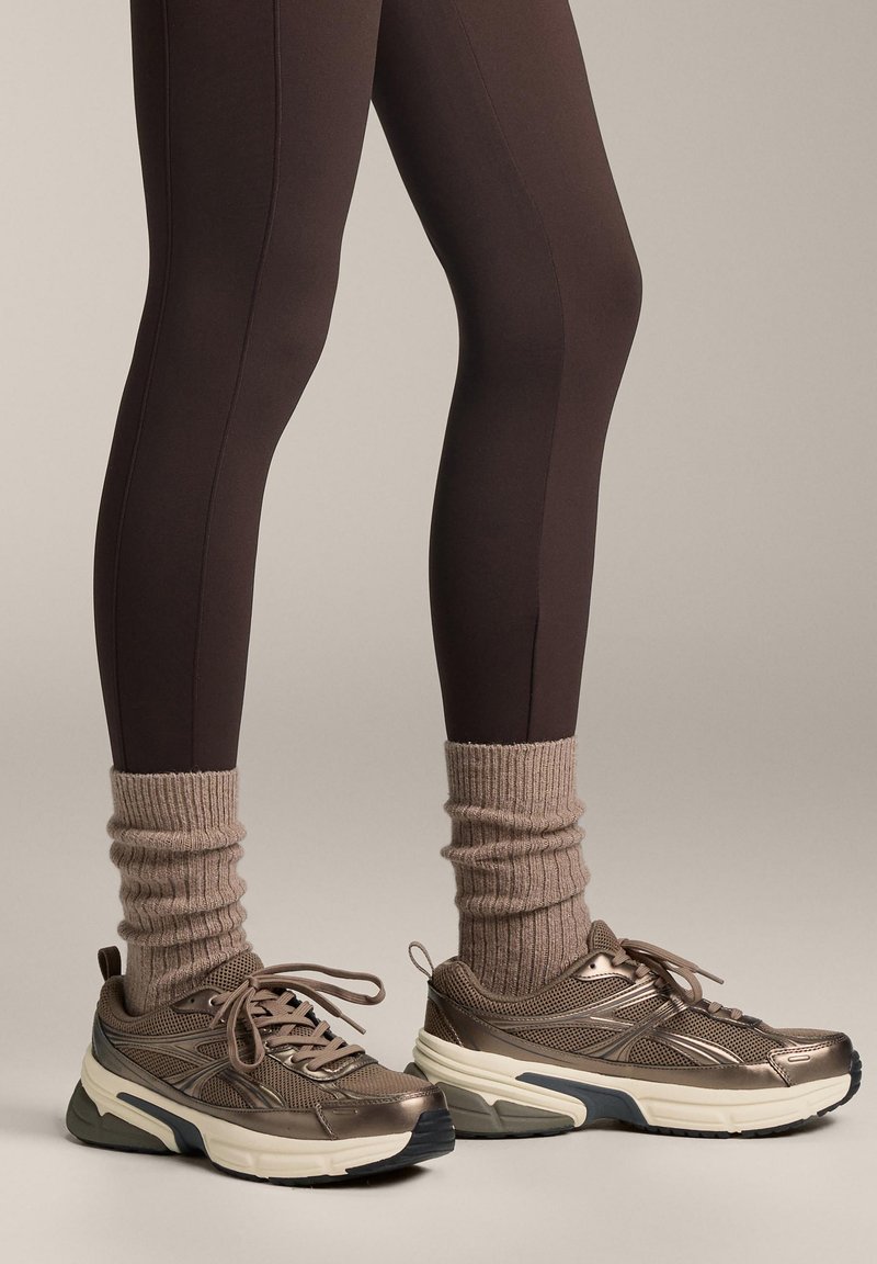 Leggings bruns avec coutures latérales, associées à des chaussettes côtelées beiges et des baskets bronze avec des panneaux en mesh et unis, semelle texturée et lacets.