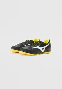 Pantofi sport negri cu talpă galbenă, având o textură matlasată, detalii albe cu logo și șireturi negre pe un interior galben.