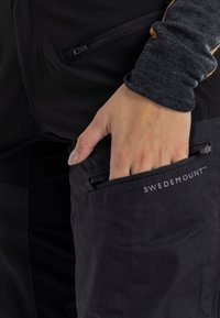 Zwarte broek met een ritszak. De stof lijkt duurzaam met een matte afwerking. Het "SWEDEMOUNT™" logo is zichtbaar nabij de bovenste zak.