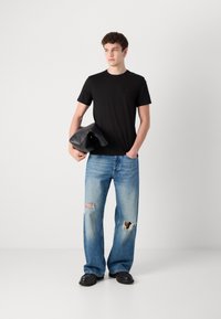 Zwarte T-shirt met korte mouwen, blauwe versleten wijde jeans met grote scheuren en zwarte leren schoenen. Model houdt een zwarte tas vast.