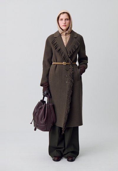 Cappotto di lana marrone con bordo frangiato e chiusura a bottone, abbinato a un top beige, pantaloni verde scuro, guanti di pelle e una borsa bordeaux.