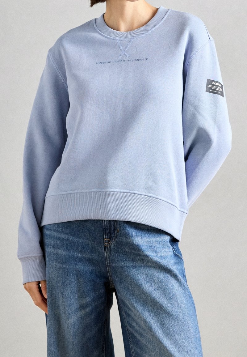 Personne portant un sweat-shirt bleu clair avec un petit texte sur la poitrine et un patch ECOALF sur la manche gauche, associé à un jean bleu.