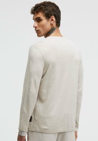 Helle beige Langarmbluse mit Rundhalsausschnitt. Weiche Textur und einfaches Design. Verfügt über ein Etikett an der unteren linken Seite.