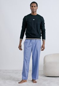 Juodas džemperis su balta logotipu ir dryžuotais rankogaliais, derinamas su šviesiai mėlynomis kelnėmis. Modelis stovintis basas ant neutralios spalvos paviršiaus.