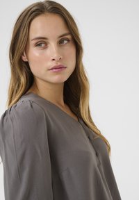 Grå blus med V-ringning, knappdetaljer, mjuk textur och volangärmar. Subtil lyster och minimalistisk design.