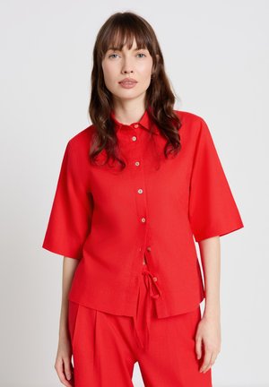 Woman with long brown hair wearing a bright red short-sleeve button-up shirt and matching red pants against a plain background.