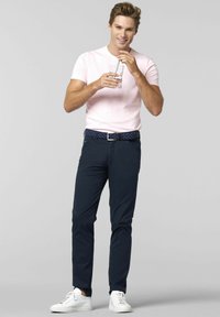 Lichtroze katoenen T-shirt, donkerblauwe broek, blauwe riem met patroon, houdt een glas met een rietje vast, witte sneakers met een gestructureerde afwerking.