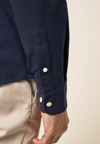 Manche d'une chemise bleu marine avec poignet boutonné et patte de boutonnage sur un pantalon beige, avec la main de la personne détendue le long du corps.