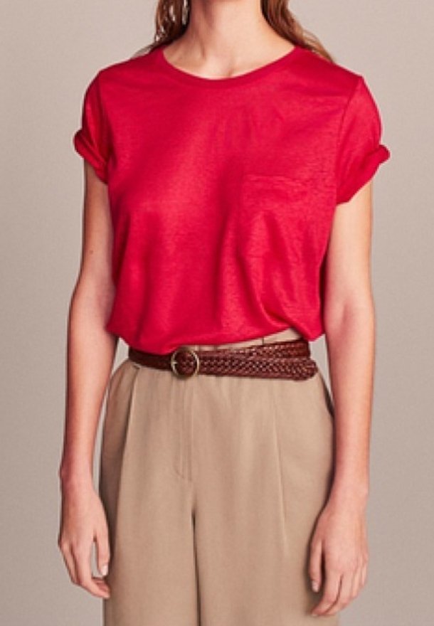 Femme porte un haut rouge vif à manches courtes avec une poche, rentré dans un pantalon beige taille haute avec une ceinture marron tressée.