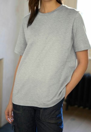 Femme portant un T-shirt à manches courtes gris clair et un pantalon en jean foncé, debout à l'intérieur avec une main dans la poche.