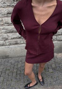 Nainen, jolla on päällään viininpunainen pitkähihainen paita ja siihen sopiva hammas sekä mustat ballerinat, seisoo mukulakivikadulla kivimuurin lähellä.