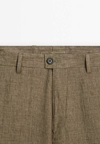 Gros plan sur la taille d'un pantalon en lin marron avec un bouton noir et des passants de ceinture sur fond blanc.