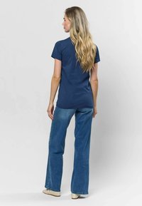 Sötétkék rövid ujjú póló, kerek nyakú, világoskék harangszárú farmerrel és bézs fonott cipővel párosítva. Hátsó nézet látható.