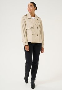 Beige dubbelzijdig trenchcoat met grote bruine knopen, brede kraag en riemlussen, gecombineerd met zwarte high-waisted broeken en enkellaarsjes.