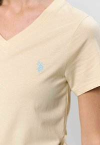 Világos bézs V-nyakú póló, puha pamutból készült, bal mellrészén világoskék hímzett logóval. Simább textúrával, rövid ujjal.