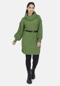 Pull vert tricoté oversize avec un col bénitier, une texture côtelée, des manches bouffantes, et une ceinture noire avec une boucle en forme de cœur, associé à des leggings noirs et des bottes de combat.