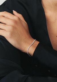 Kéz finom gyémánt teniszkarkötővel, amely az ellentétes vállon pihen, fekete hosszú ujjú ruhát viselve.