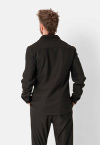 Zwarte, geribbelde shirt met lange mouwen en een kraag, voorzien van knopen op de manchetten en een rechte achterzoom. Het model wordt van achteren weergegeven.