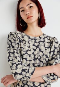 Haljina s cvjetnim uzorkom, nabrana u krem i tamnoplavu boju. Ima okrugli izrez i rukave do lakta s dugmadi na krajevima. Teksturirani materijal.