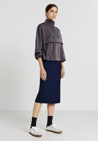Grau pullover mit hohem Kragen und Taschen auf der Vorderseite, kombiniert mit einem marineblauen Bleistiftrock. Weibliches Model trägt weiße Sneaker und schwarze Socken.