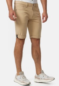 Tan, knälånga shorts i bomull med knapp och dragkedja fram. Rullade fållar med svart hårdvara. Bärs med grå sneakers.