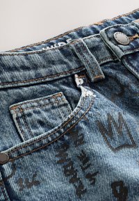 Jeans in denim blu presentano un tessuto strutturato con finitura consumata, stampe decorative nere e cuciture arancioni a contrasto sulla vita.