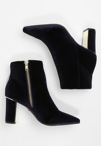 Botas de tobillo de terciopelo azul marino con puntera afilada, detalle de cremallera dorada y tacón robusto con acento metálico. Textura lisa, diseño elegante.