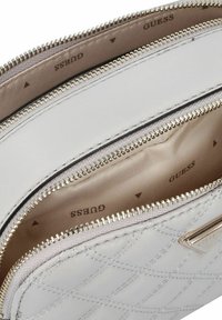 Portefeuille en cuir matelassé blanc avec des zips dorés et une doublure beige. Dispose de plusieurs compartiments pour une meilleure organisation. Logo visible à l'intérieur.