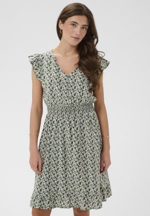 Mladá žena s dlhými hnedými vlasmi, oblečená v zelenej kvetinovej šaty dĺžky po kolená s volánmi na rukávoch a so žabičkovaným pásom, stojaca pred svetlým pozadím.