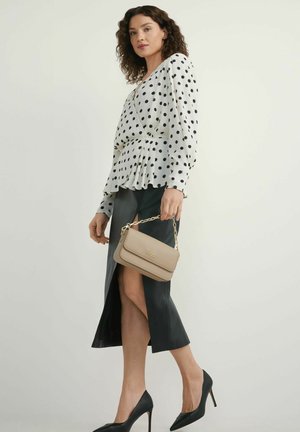 Kobieta w białej bluzce w czarne kropki, czarnej spódnicy z rozcięciem, czarnych szpilkach, trzymająca beżową torebkę z złotym łańcuszkiem.