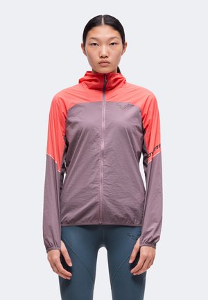 Donna che indossa una giacca leggera con zip con spalle e maniche corallo, corpo viola e leggings blu scuro, in piedi rivolta in avanti.