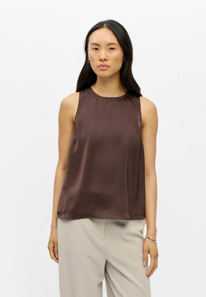 Femme aux longs cheveux foncés porte un haut marron sans manches et un pantalon beige clair, debout devant un fond blanc uni.