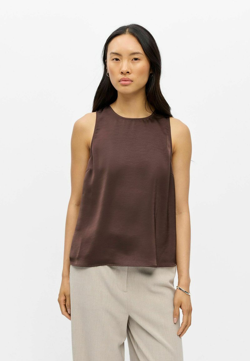 Femme aux longs cheveux foncés porte un haut marron sans manches et un pantalon beige clair, debout devant un fond blanc uni.