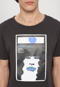 Donkergrijze katoenen t-shirt met een gedrukt grafisch ontwerp van een gezicht met blauwe lippen en ogen, overlappend met tekst in blauw en wit, en een blauw hart.