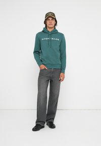 Felpa verde con logo bianco, cordini e tasca a marsupio, abbinata a jeans grigi a gamba larga e scarpe nere. Postura eretta e sfondo pulito.