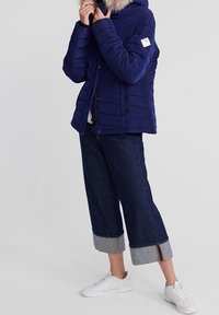Marineblå pufferjakke med en hætte fyldt med pels, lynlås og quiltet design. Matchet med brede mørkeblå denimbukser med ophøjede sømme.