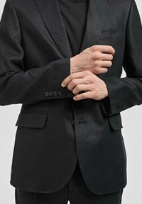Schwarzer Blazer aus strukturiertem Stoff, einreihiger Schnitt, mit Reverskragen und vier Knopfdetails an den Ärmeln. Maßgeschneiderte Passform.