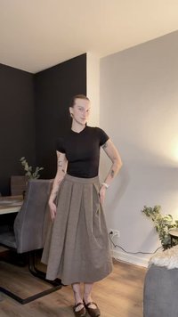 Bruine geplooide rok met een gestructureerde afwerking, gecombineerd met een zwart aansluitend topje. Bruine schoenen en een neutrale binnenomgeving met planten en meubels.