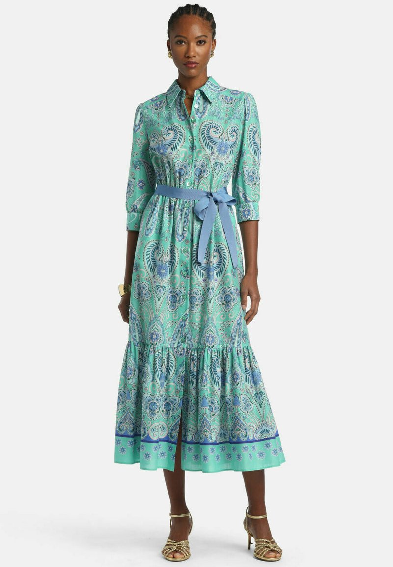 Woman wearing a long mint green dress with blue paisley patterns, a blue waist tie, and gold high-heeled sandals, standing against a white background.