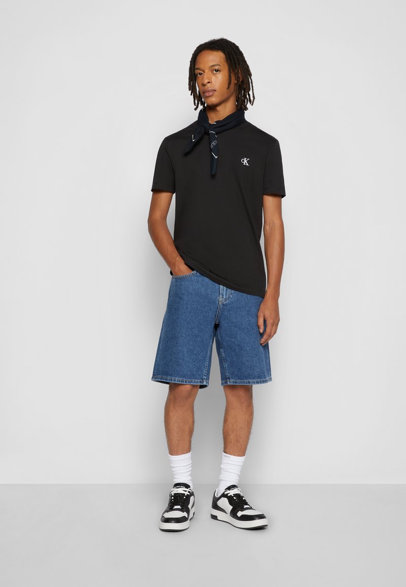 Joven con rastas que lleva una camiseta negra de manga corta, pantalones cortos vaqueros azules, zapatillas negras y blancas, calcetines blancos y un pañuelo negro en el cuello.